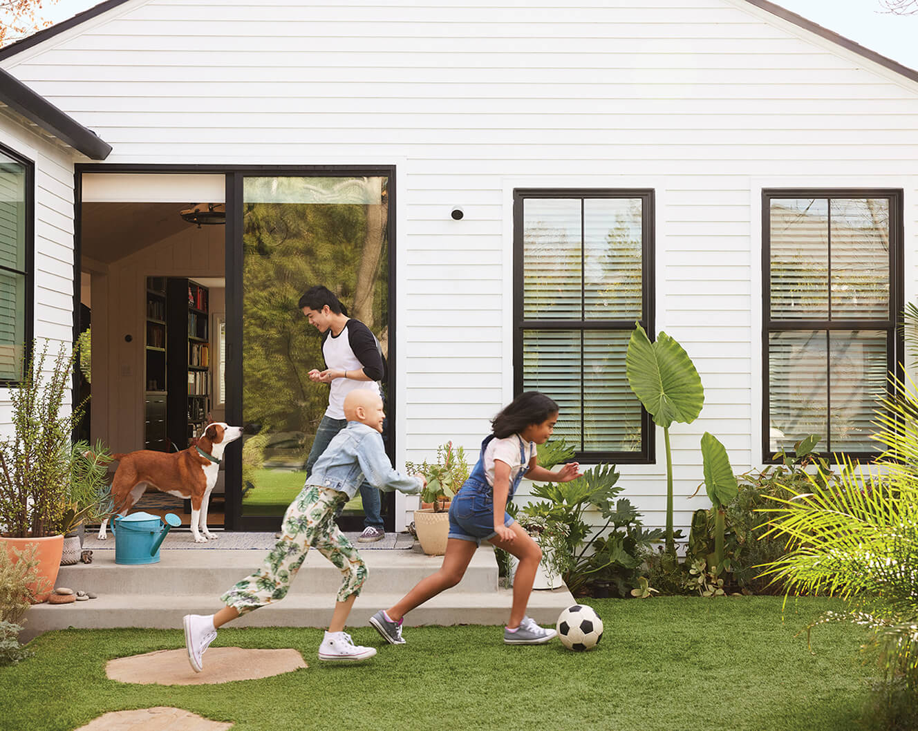 Two children play soccer in a backyard while an adult and a dog stand near an open door to the house. A Google Nest Cam (outdoor or indoor, battery) is installed on the side of the house.